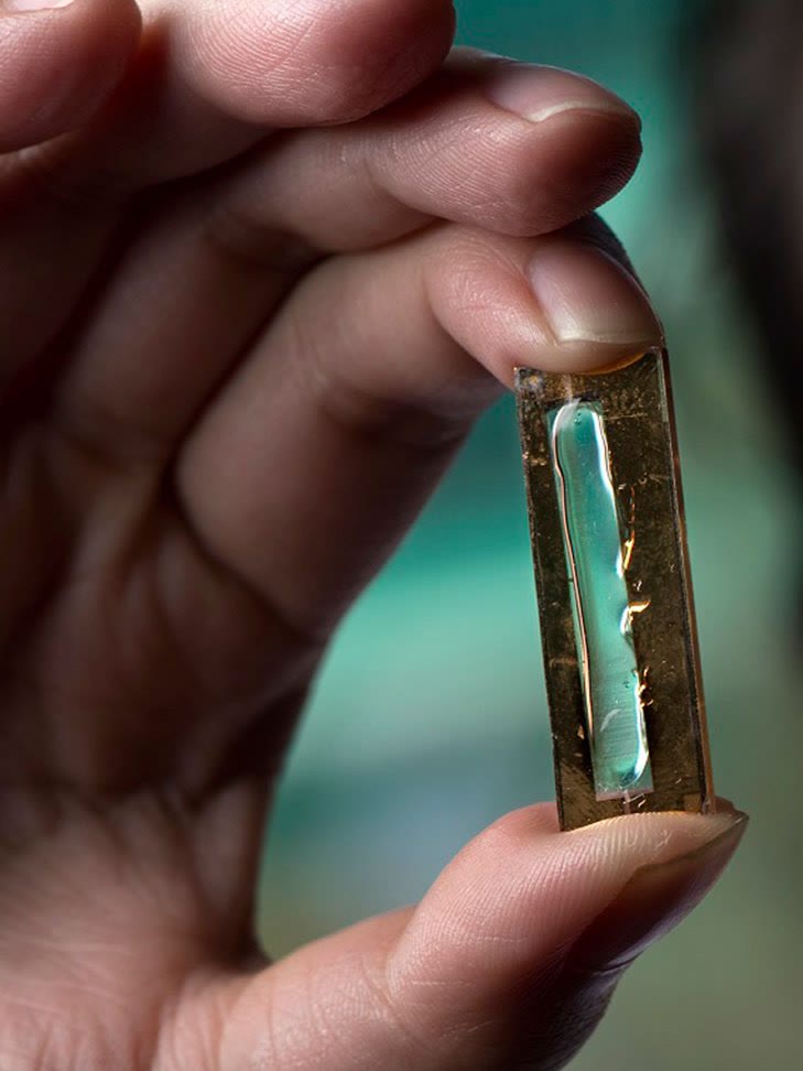 Fingers hold a small gold-framed cell with a bright teal strip inside.