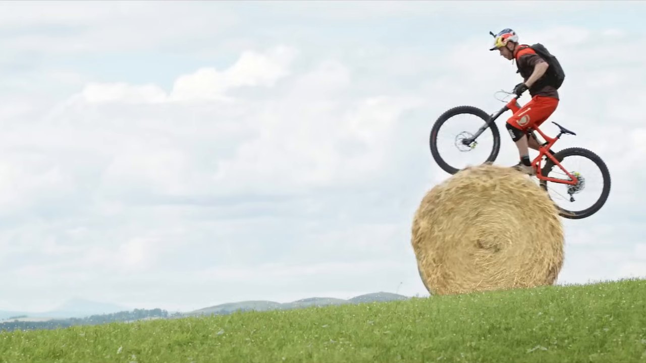 A mountain biker in red balancing his bike on top of a round hay bale on a grassy hillside.