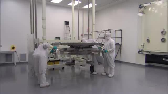 People in white cleanroom suits around a large round mirror on a wheeled stand.