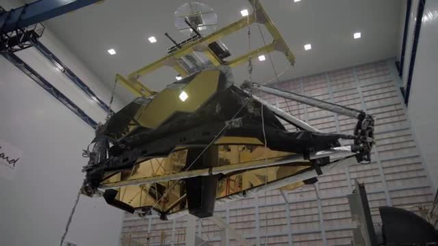 The gold-and-black Webb telescope hanging from a yellow crane inside a tall white hall.