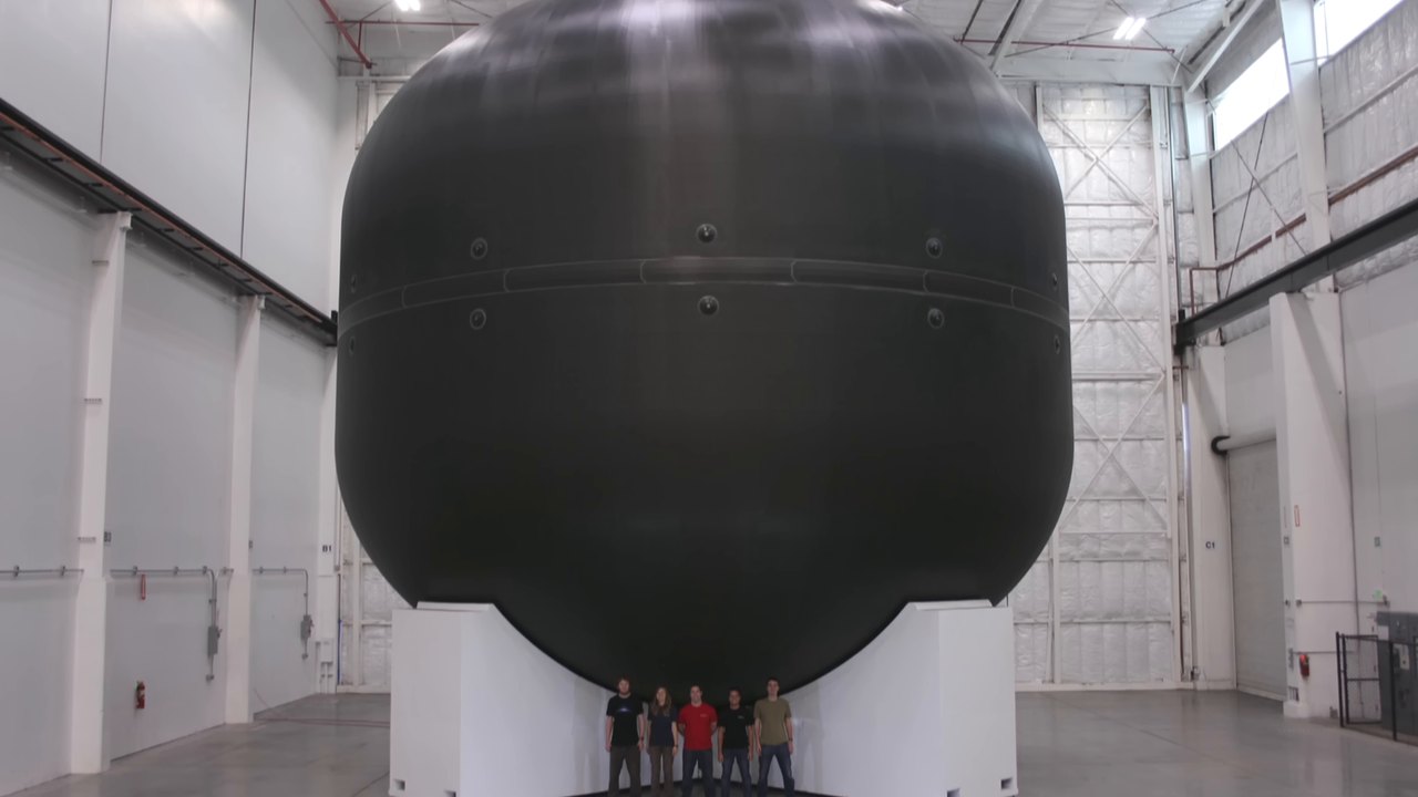 A huge black carbon-fibre propellant tank on a white stand in a factory, with five people standing beneath it.