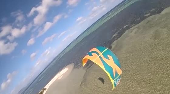 First-person view of a teal and orange kite flying low over sand toward a turquoise lagoon.