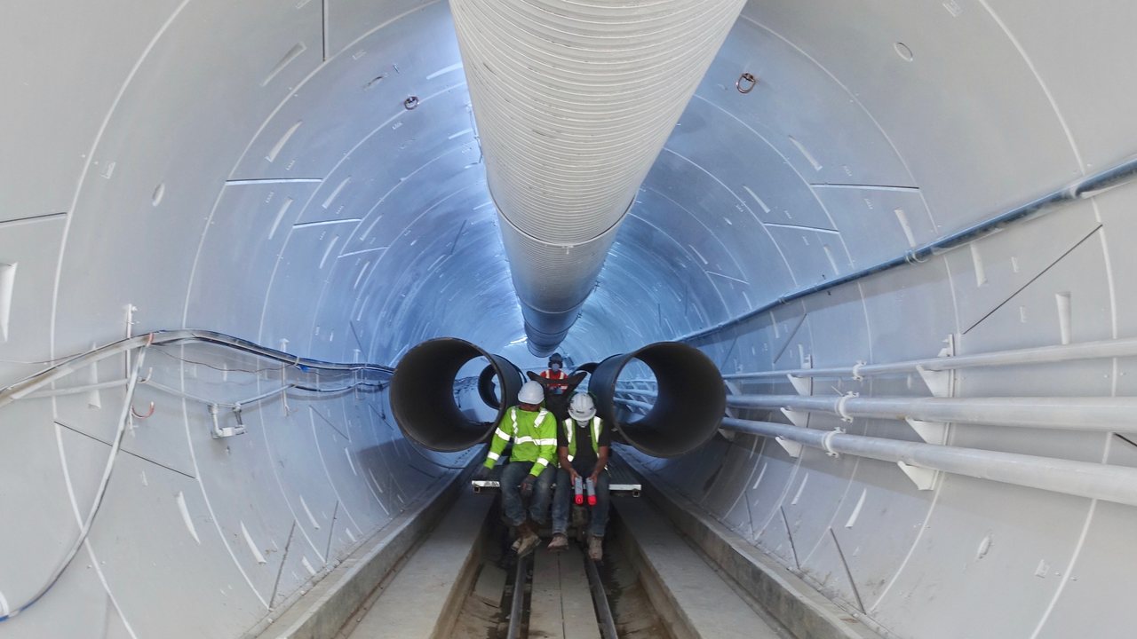 Two workers in hard hats ride a rail sled through a long, round Boring Company test tunnel in Hawthorne, California.