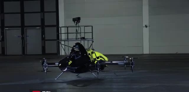 A person seated in a black and yellow multirotor craft inside a hangar