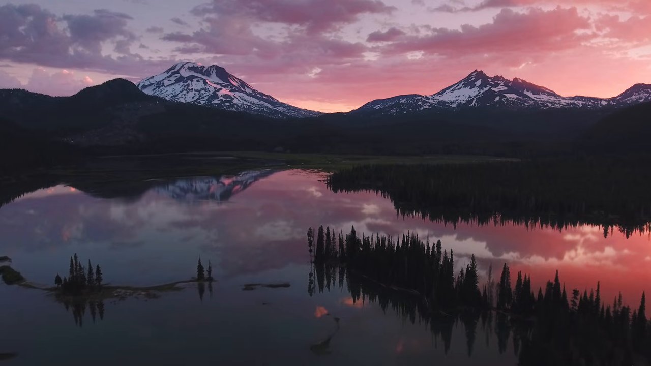 Snow-capped Cascade peaks and dark forest reflected in a still Oregon lake under a pink sunset sky.
