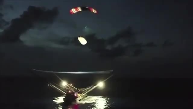 A Falcon fairing half under a parachute descending toward a lit catch net at sea