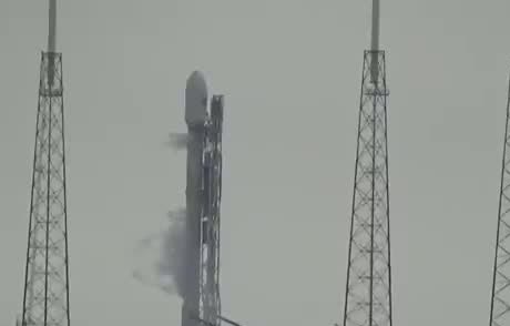 A rocket on the pad between lightning towers, with smoke at its base.