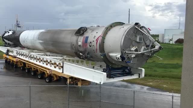 A used Falcon 9 booster lying on a transporter behind a fence.