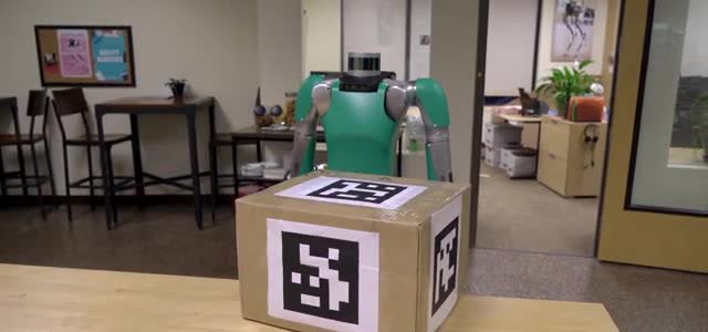 A green and silver humanoid robot standing behind a cardboard box with black-and-white markers.
