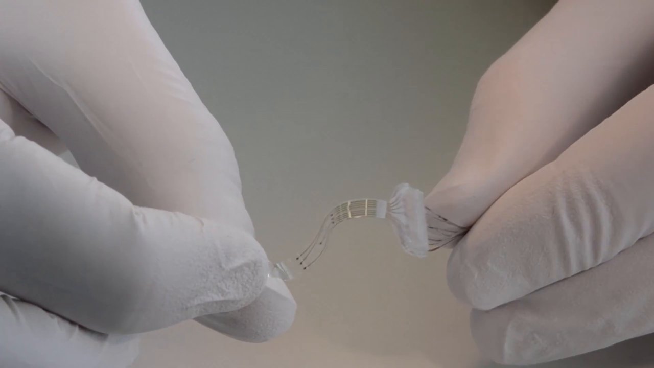 Gloved fingers stretching a thin, transparent e-dura spinal implant with fine electrode tracks.