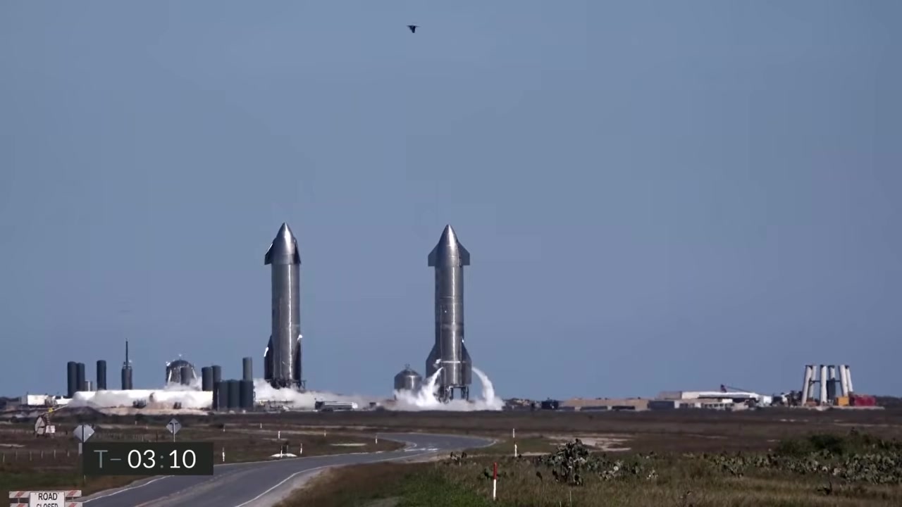 Two Starship prototypes venting vapor on pads under a blue sky