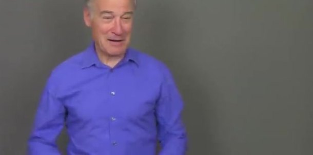 A man in a purple shirt against a plain grey wall, generated rather than filmed.