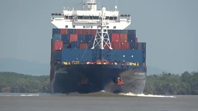 A container ship seen head-on, pushing a bow wave along the river.