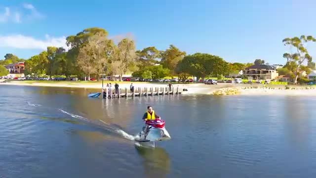A small foiling craft skimming across calm water near a tree-lined shore.