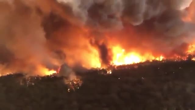 A bushfire front burning through scrub under a sky filled with orange smoke.